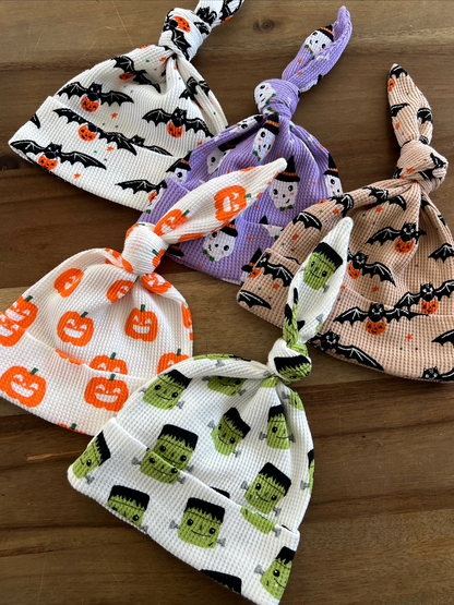 Four Halloween-themed knotted baby hats featuring pumpkins, ghosts, and Frankenstein characters on a wooden surface.