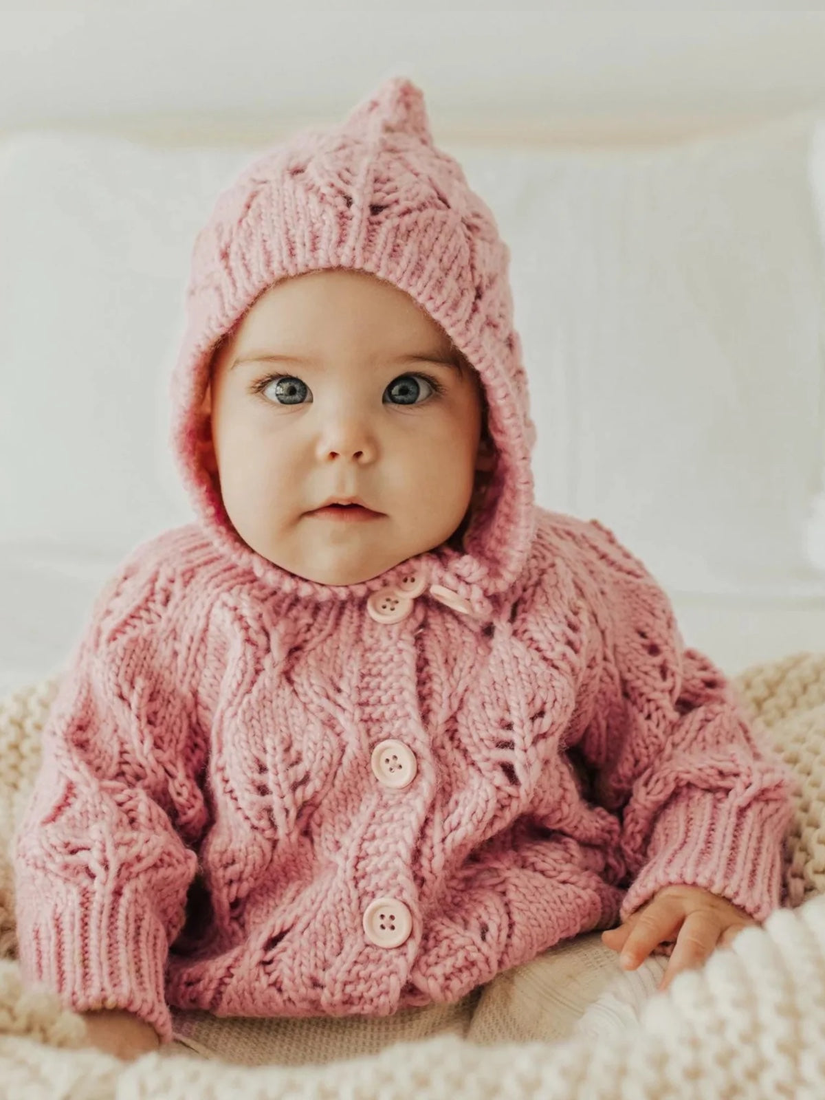 Baby in a cozy pink knitted cardigan and hat, sitting on a soft blanket with an adorable expression.