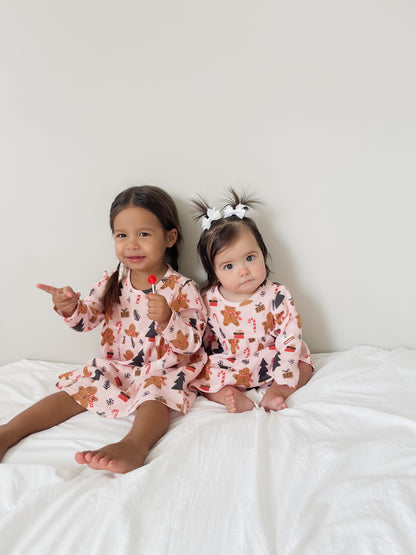 Two toddlers in matching gingerbread-patterned dresses sit on a white blanket, smiling and playing with candy.