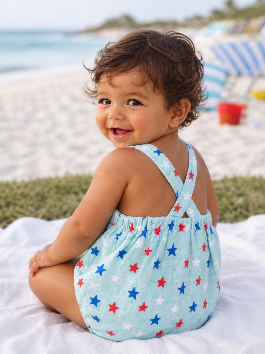 Smiling toddler in a stars-patterned outfit sits on a towel by the beach, with ocean waves in the background.