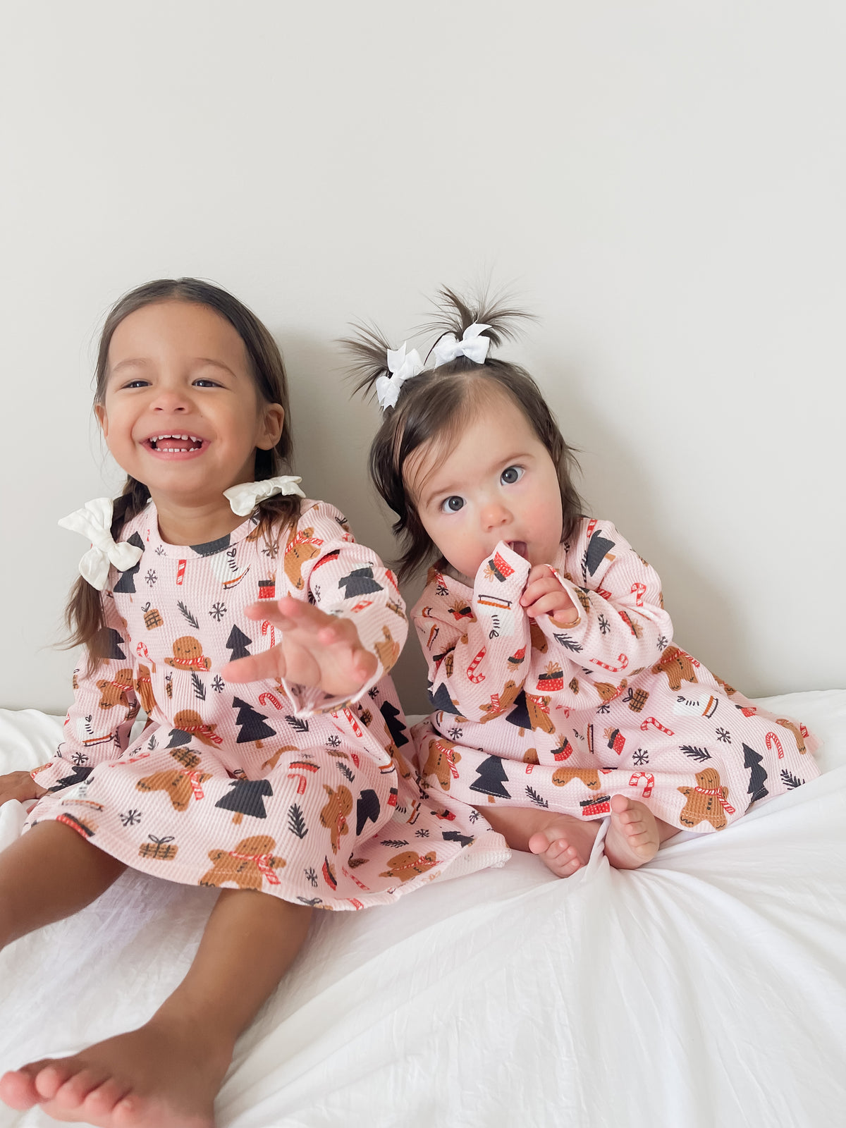 Two young girls in festive dresses sit on a white bed, smiling and playing with playful expressions.