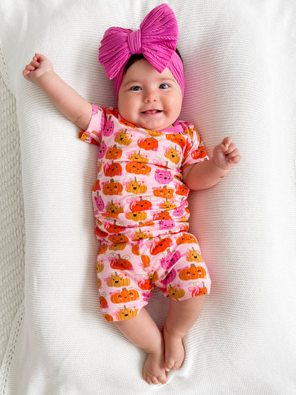 Smiling baby in a pumpkin-patterned outfit with a large pink bow, lying on a soft white blanket.