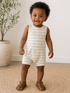 Smiling toddler in striped romper stands on a jute rug, with a potted plant in the background.