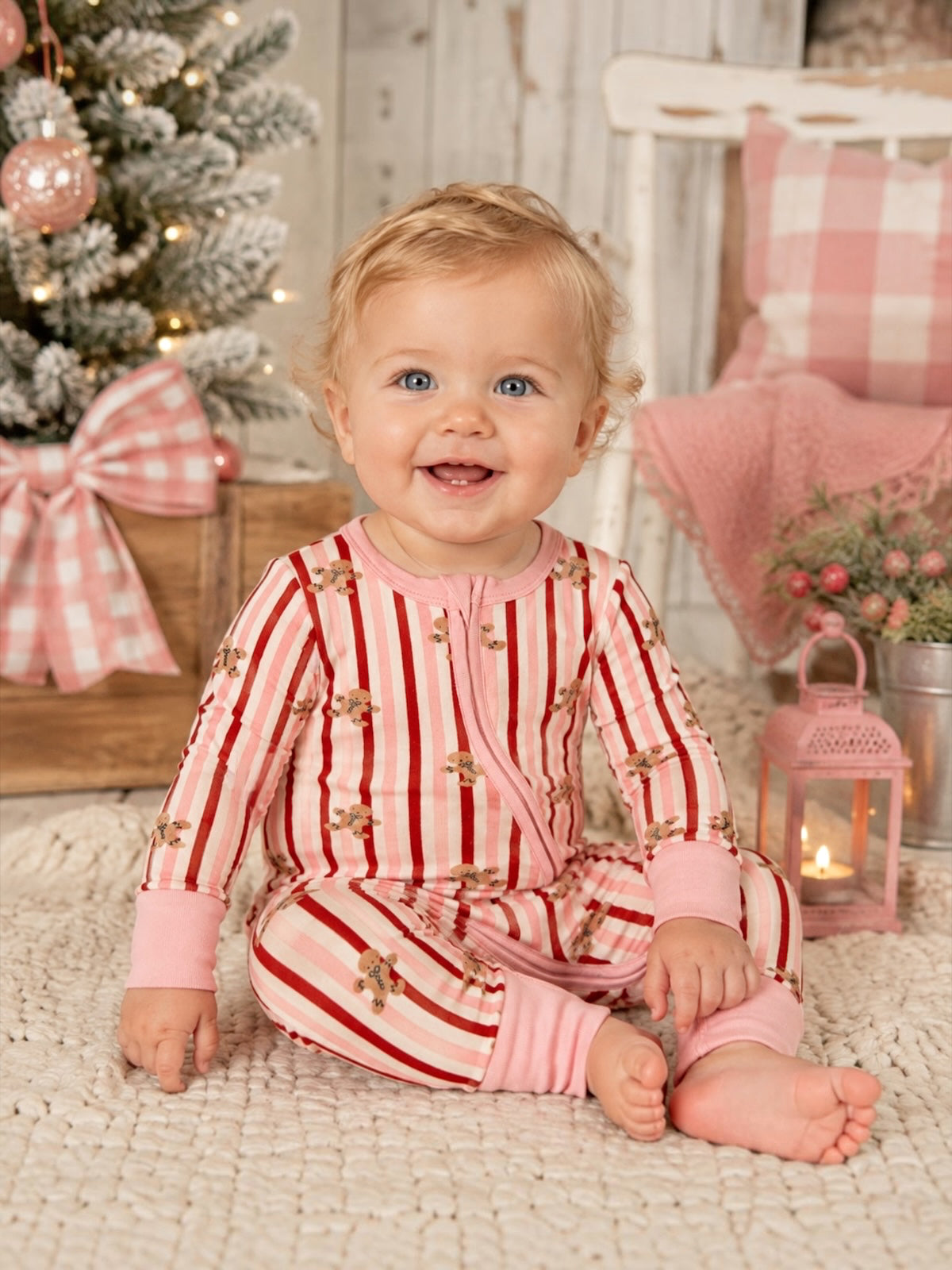 Smiling baby in red and pink striped pajamas sitting on a cozy rug, with a decorated Christmas tree in the background.