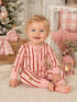Smiling baby in red and pink striped pajamas sitting on a cozy rug, with a decorated Christmas tree in the background.