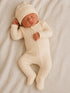 Newborn baby sleeping in a cream-colored ribbed outfit and matching hat, resting on a textured blanket.
