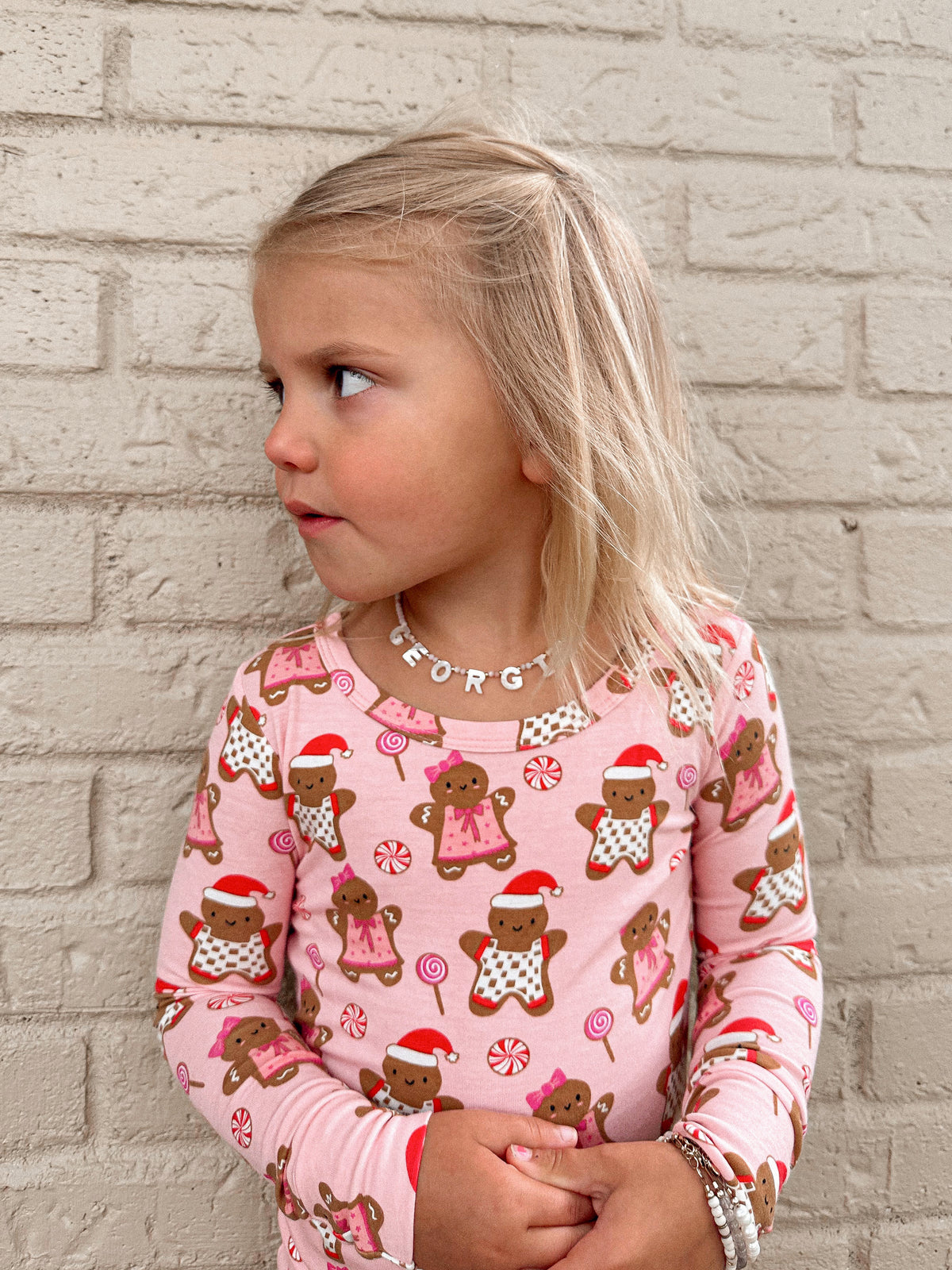 Young girl in a pink gingerbread-patterned shirt, looking sideways against a brick wall background.