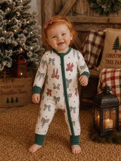 Toddler in festive pajamas with bows, smiling in a cozy holiday setting with a Christmas tree and lantern.