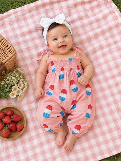 Smiling baby on a picnic blanket, wearing a pink outfit with popsicle prints, surrounded by strawberries and flowers.