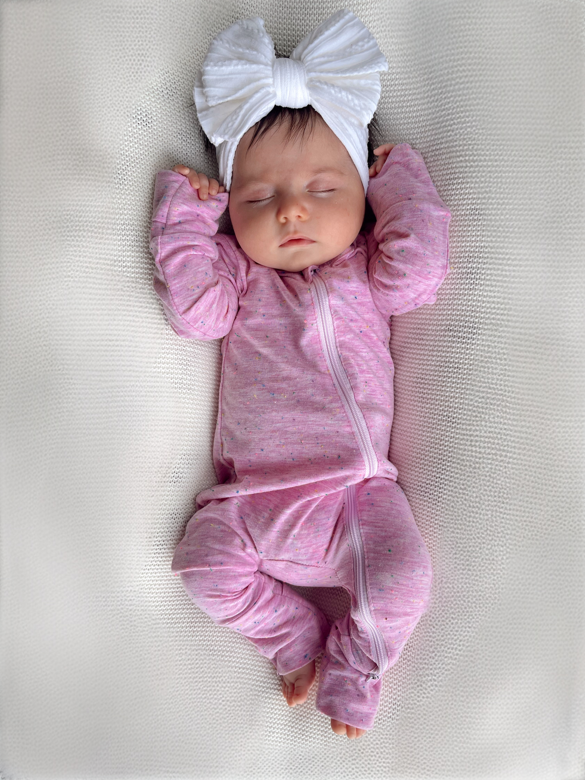 Sleeping baby girl in pink onesie with white bow headband, resting on a textured blanket.