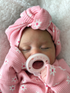 Baby sleeping peacefully with a pink headband and pacifier, wrapped in a cozy pink blanket with floral patterns.