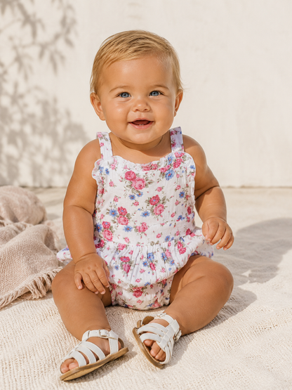 Smiling baby in floral romper sits on sand, wearing white sandals, with a bright, sunny background.