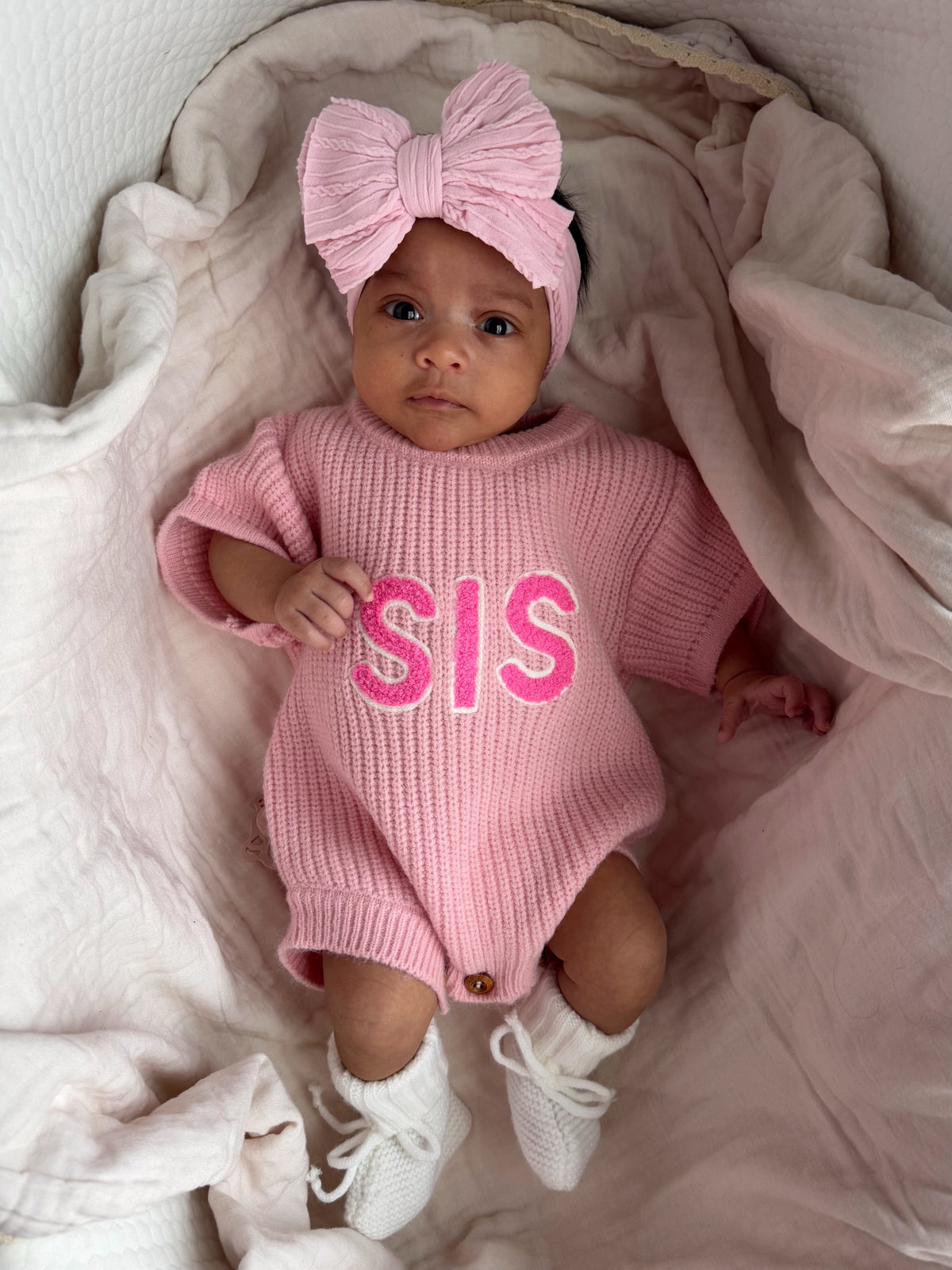 Baby girl in a pink knit outfit with "SIS" text, wearing a matching bow headband and white booties, lying on soft fabric.