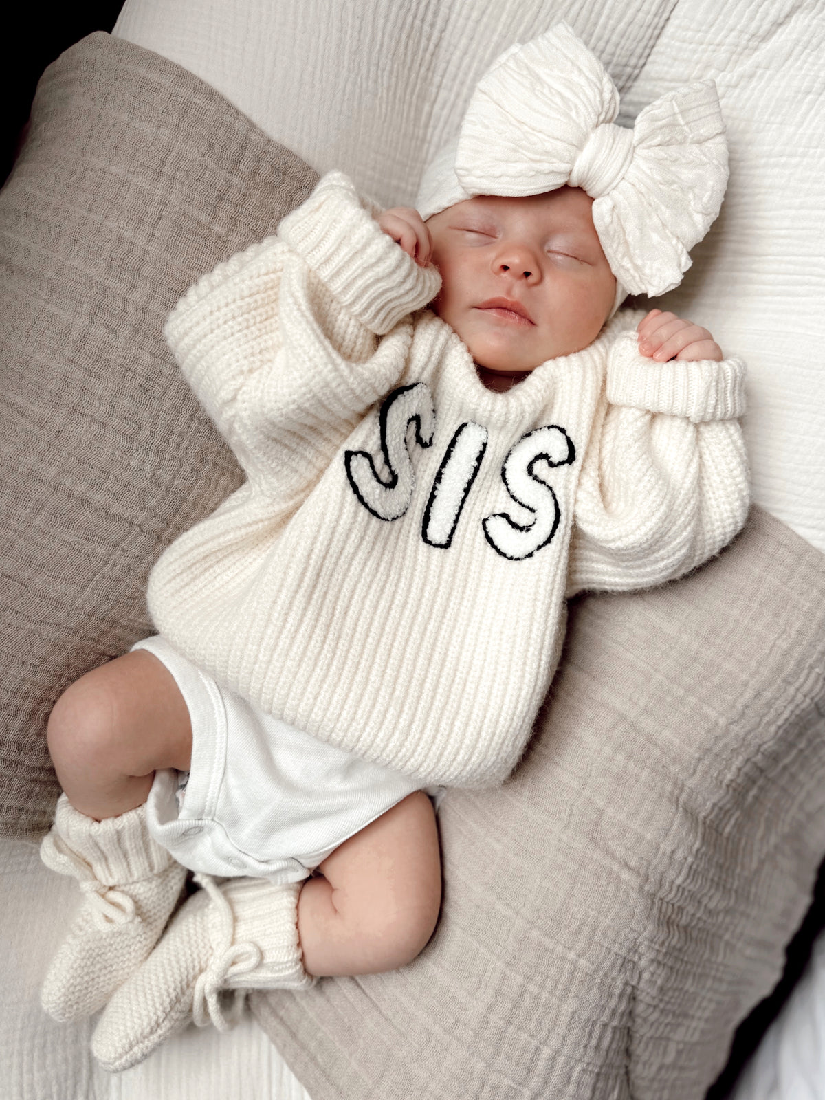 Baby wearing a cozy white sweater with "SIS" printed, resting on soft pillows with a large bow headband.