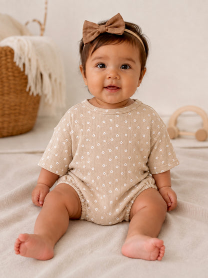 Smiling baby in a beige outfit with floral prints and a brown bow, sitting on a soft textured surface.