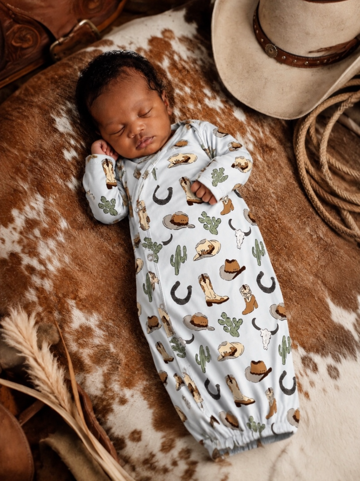 Sleeping baby in cowboy-themed onesie, nestled on a patterned blanket with a hat and cowboy accessories nearby.