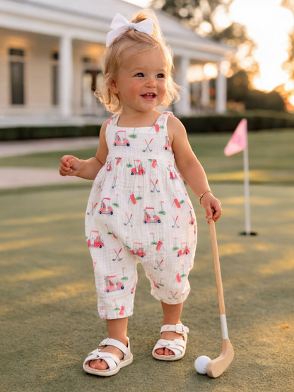 Cute toddler in a white outfit with golf patterns, standing on a putting green, holding a golf club and smiling.