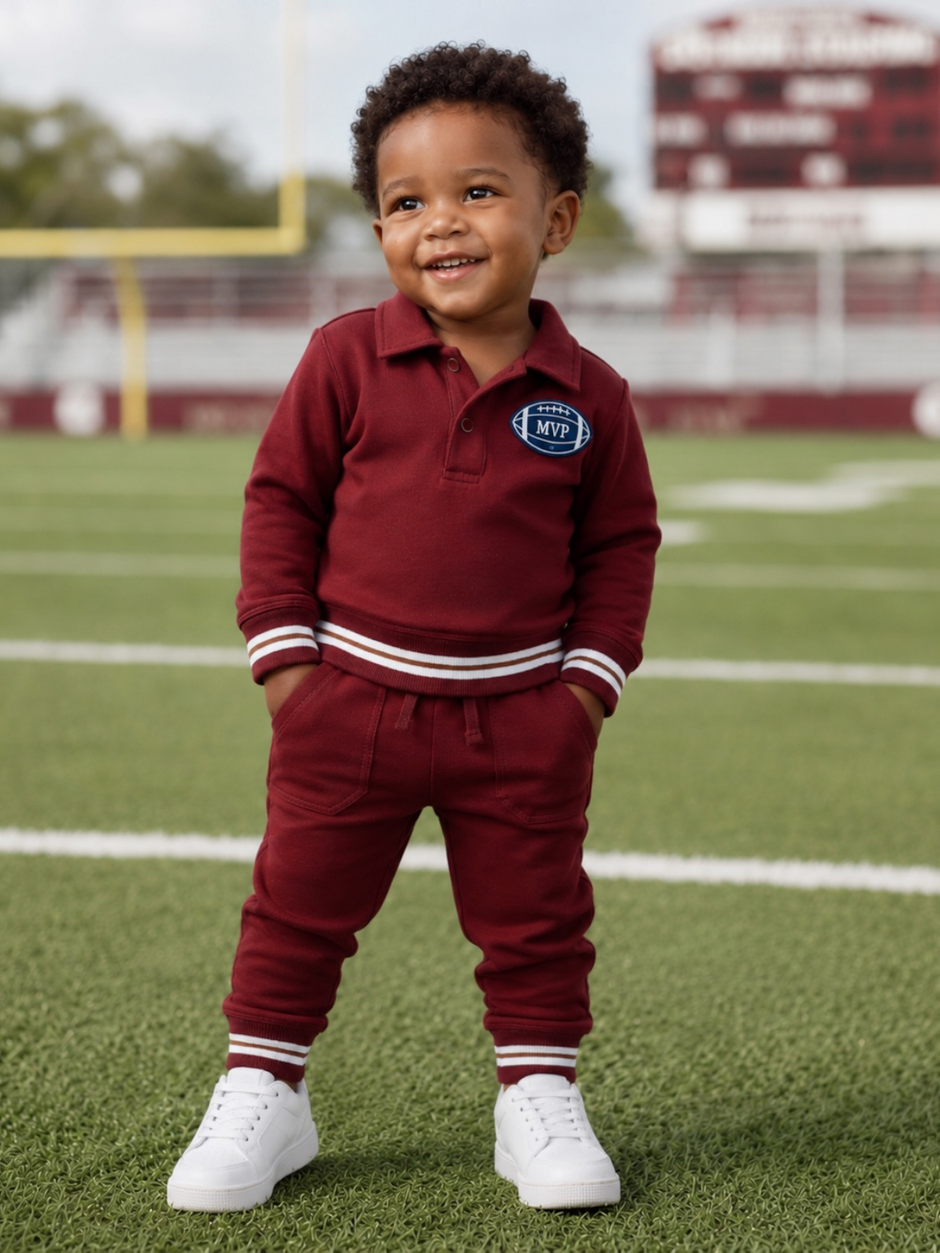 Smiling toddler in maroon outfit stands on a football field, wearing white sneakers and an MVP patch.