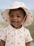 Smiling child in a hat and palm-patterned shirt, with a beach in the background.
