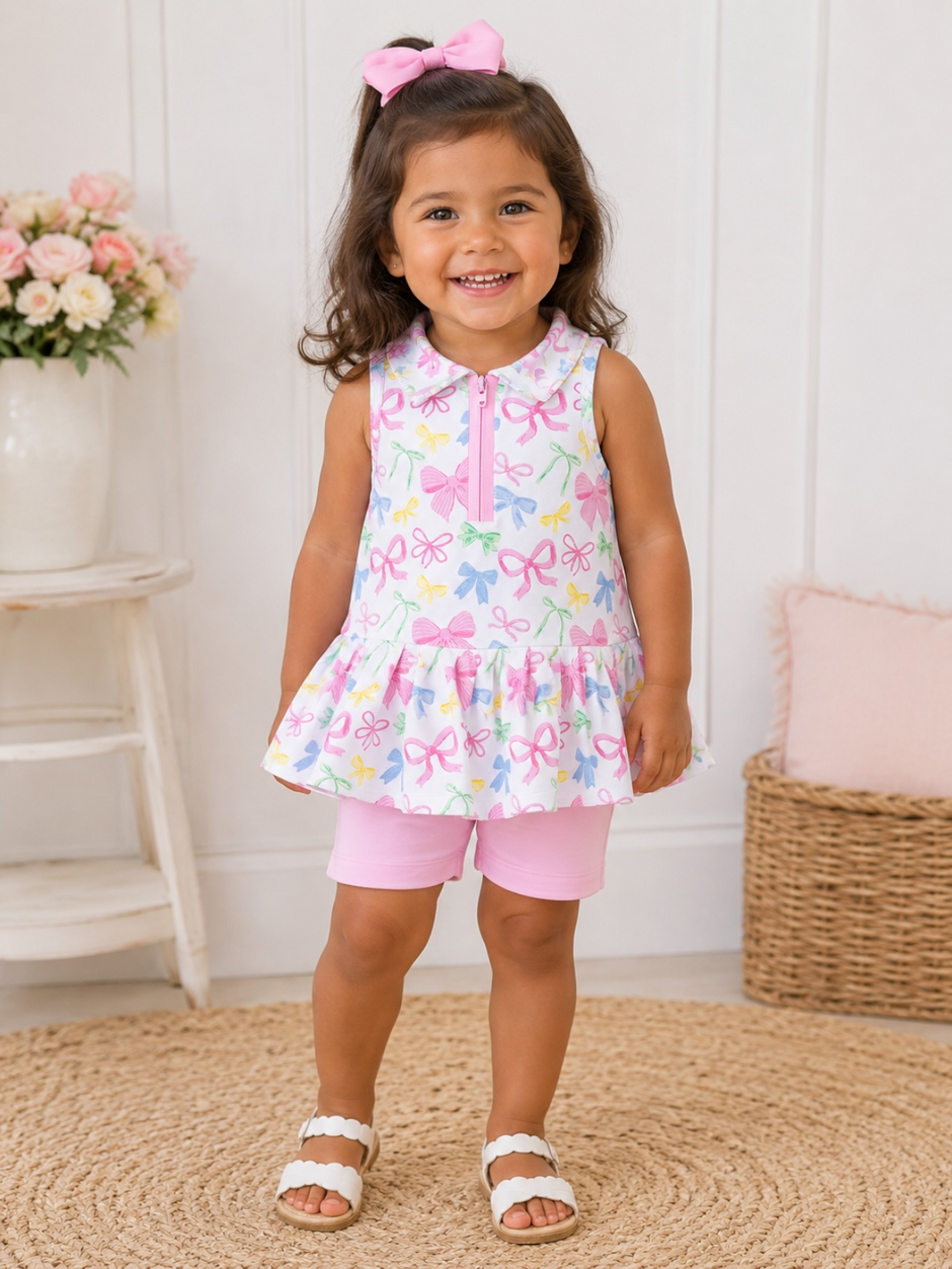 Smiling young girl in a colorful dress and pink shorts, standing on a woven rug with flowers in a vase nearby.
