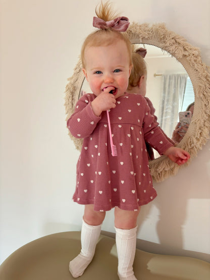 Smiling toddler in a heart-patterned dress holding a pink toothbrush, standing by a decorative mirror.
