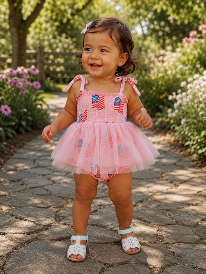 Smiling toddler in a pink dress with tulle, standing on a stone path surrounded by flowers and greenery.