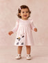 Smiling toddler in a pink striped dress with a cow design, wearing a hair bow and white shoes, standing on a pastel background.
