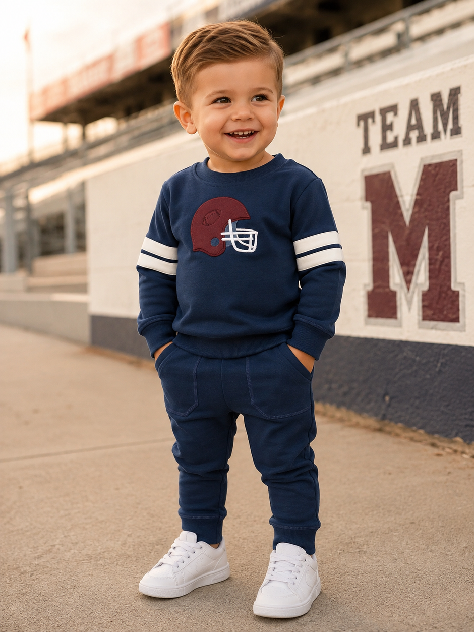 Smiling toddler in navy outfit with football design, standing on pavement near stadium, wearing white sneakers.