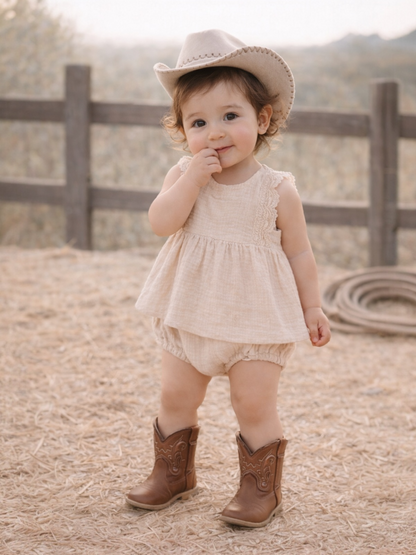 Toddler in a beige dress and cowboy hat, smiling while standing on straw-covered ground near a wooden fence.
