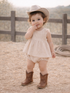 Toddler in a beige dress and cowboy hat, smiling while standing on straw-covered ground near a wooden fence.