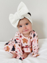 Smiling baby girl in a festive outfit and large white bow, sitting on a white blanket.