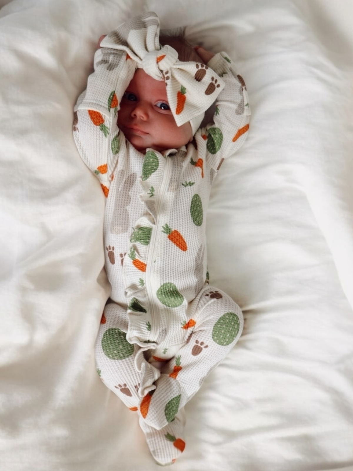 Infant laying on soft fabric, wearing a patterned onesie with carrots and cacti, adorned with a large bow.