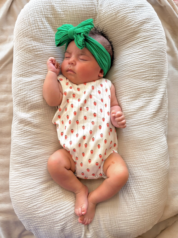 Baby sleeping on a soft surface, wearing a white outfit with red strawberries and a green headband.