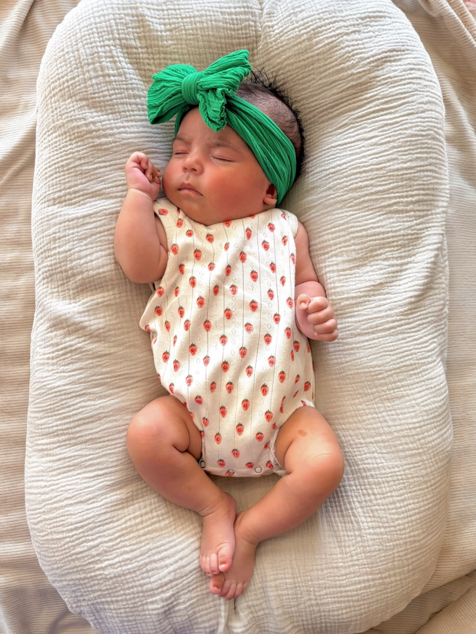 Baby sleeping on a soft surface, wearing a white outfit with red strawberries and a green headband.