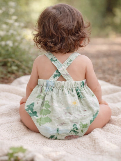 A toddler with curly hair sits on a blanket, wearing a floral romper in a forest setting.