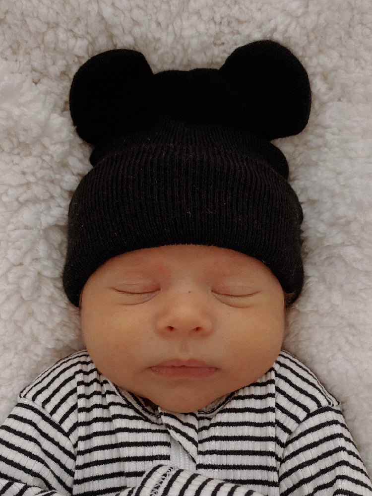Sleeping baby wearing a black bear-ear beanie on a soft white blanket, dressed in black and white stripes.