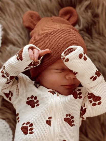 Newborn baby in a cozy, paw-print onesie and a brown bear-ear hat, peacefully resting on a soft blanket.