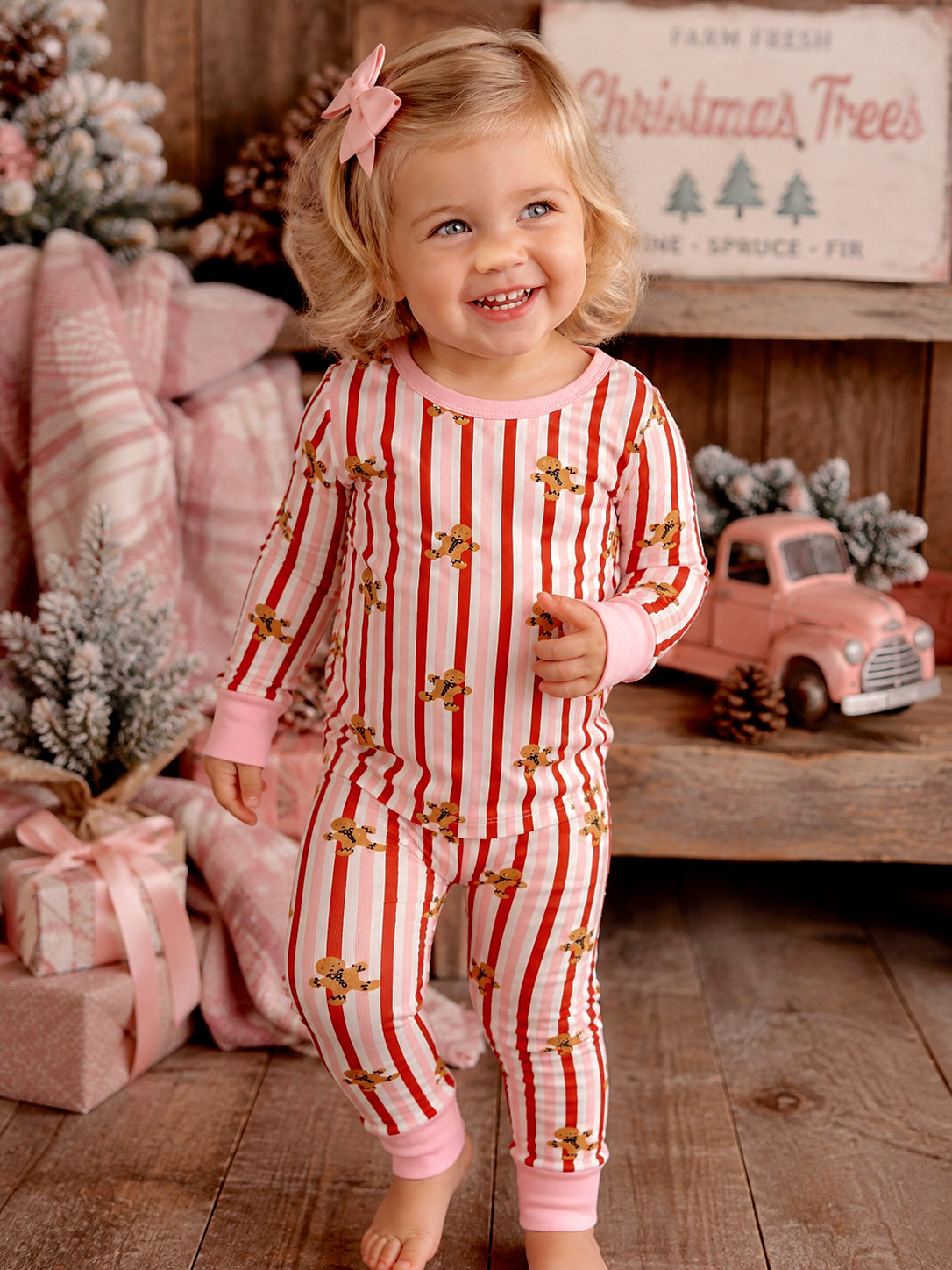 Smiling toddler in striped pajamas with gingerbread prints, surrounded by holiday decor and gifts.