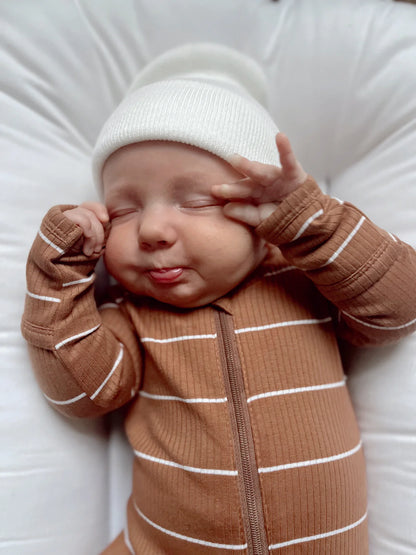Baby in brown striped outfit and white hat making a playful face, sticking out tongue and squinting eyes.