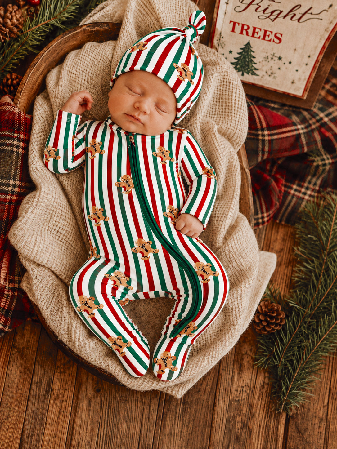 Sleeping baby in festive striped outfit with gingerbread design, nestled in a cozy basket surrounded by holiday decor.