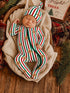 Sleeping baby in festive striped outfit with gingerbread design, nestled in a cozy basket surrounded by holiday decor.