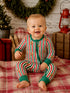 Smiling baby in festive striped pajamas, sitting on a blanket with holiday decor in the background.