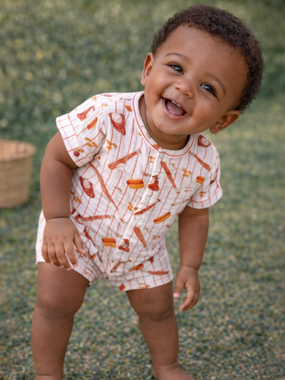 Smiling baby in a playful outfit, standing on grass with a woven basket in the background.