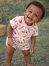Smiling baby in a playful outfit, standing on grass with a woven basket in the background.