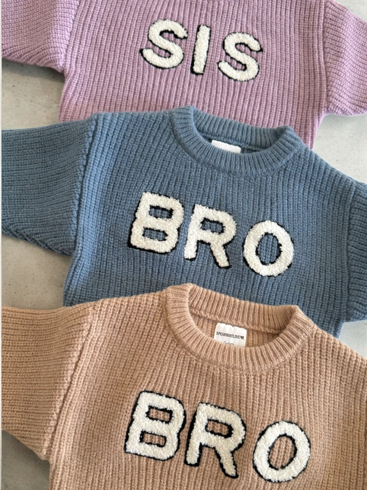 Three knitted sweaters in pastel colors with the words "SIS" and "BRO" prominently displayed.