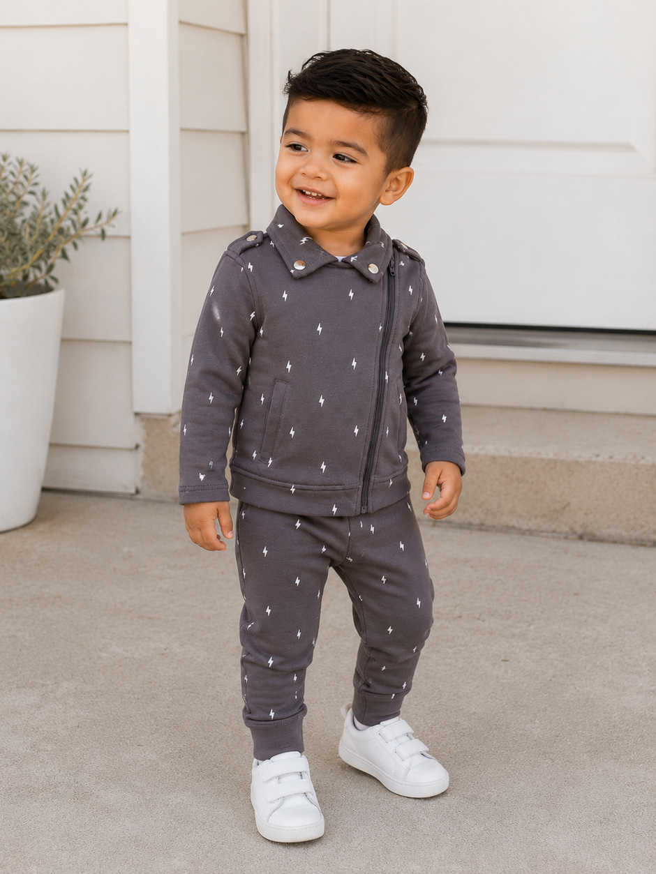 Smiling young boy in a gray outfit with lightning patterns, standing on a porch, wearing white sneakers.