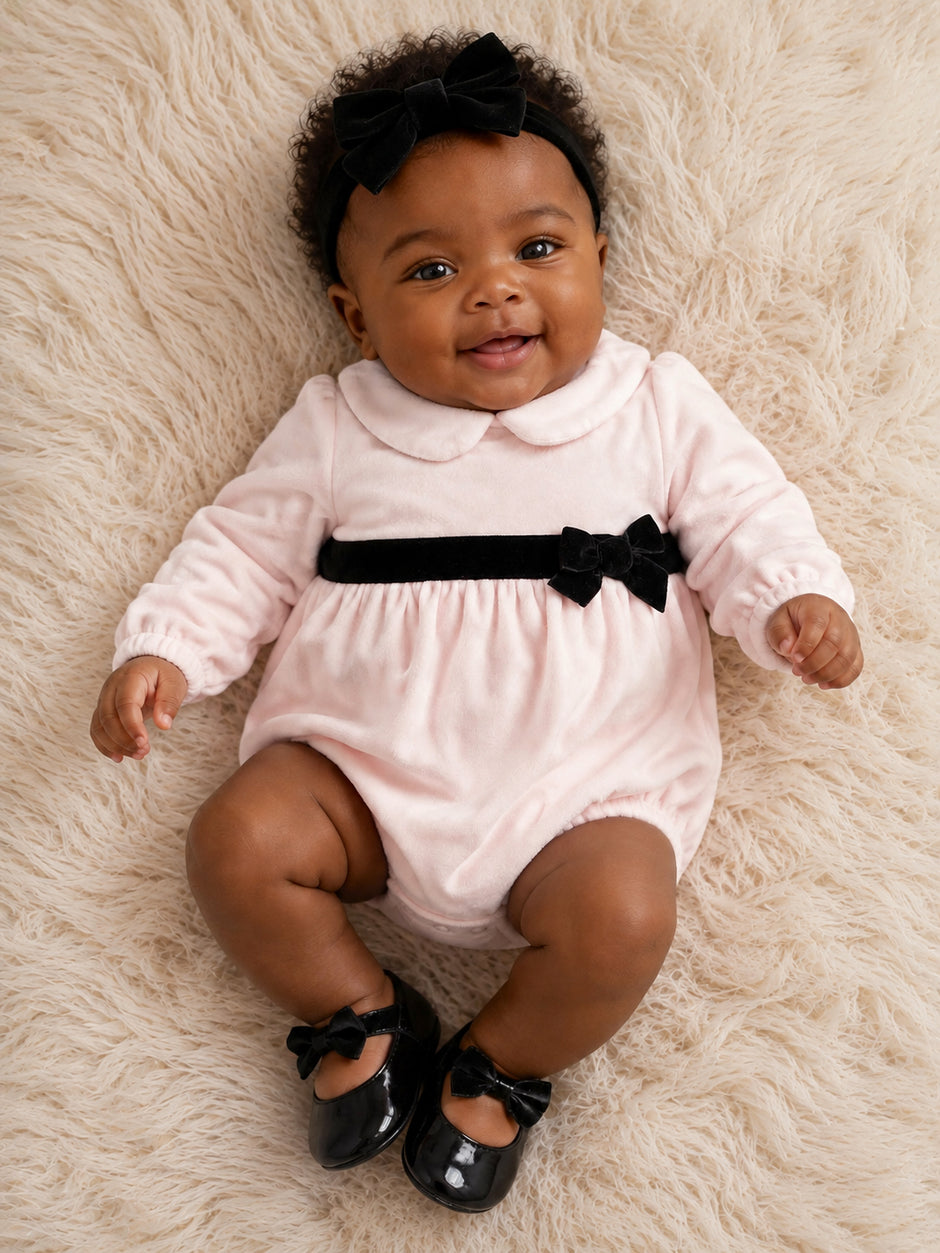 Smiling baby in a pink romper with a black bow on a soft, textured surface.