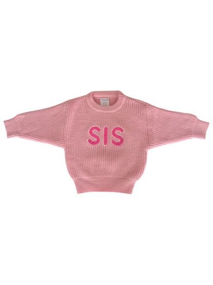 Pink knitted sweater with "SIS" printed across the front, featuring wide sleeves and a ribbed collar.
