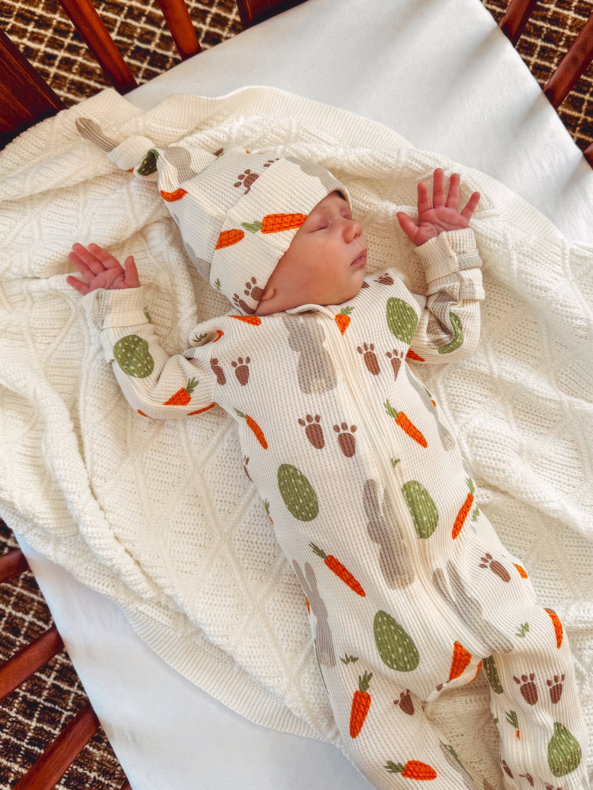 Infant sleeping on a textured blanket, wearing a patterned onesie with carrots and animals, in a cozy setting.
