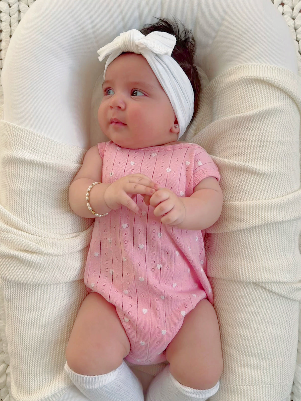 Baby in a pink outfit with a white headband, sitting on a soft, textured surface, looking thoughtfully to the side.